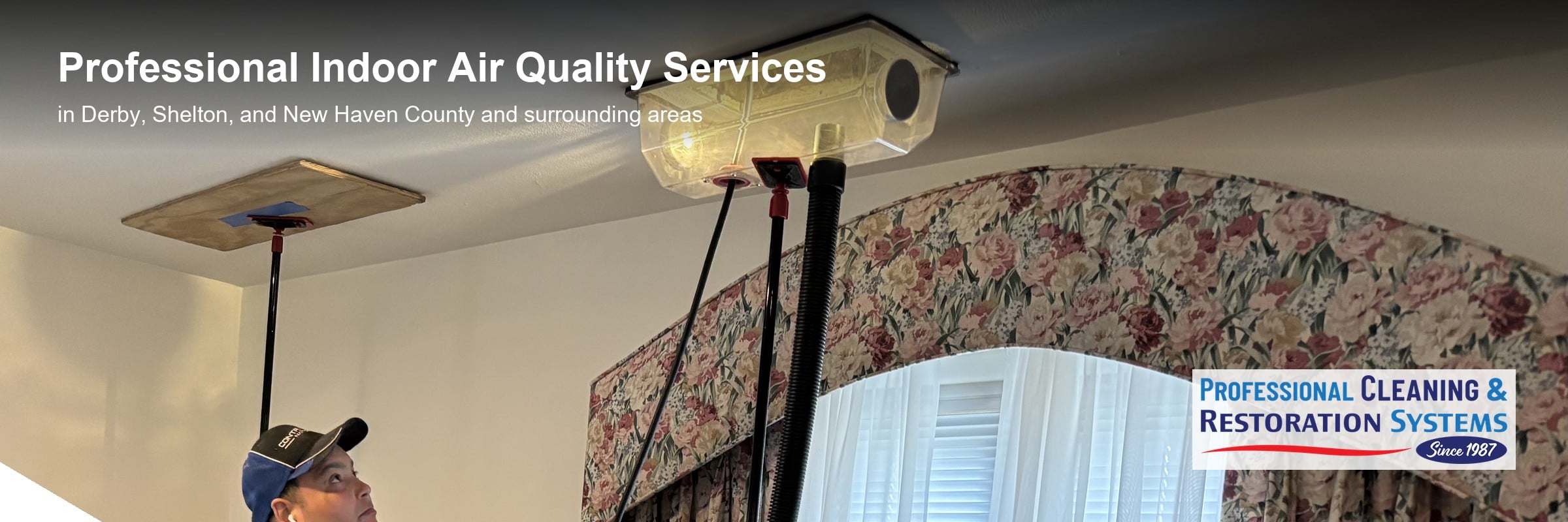 Indoor air quality work in Derby CT — a PCRS technician working a ceiling supply register with a vacuum line and a work light, a second register open across the room