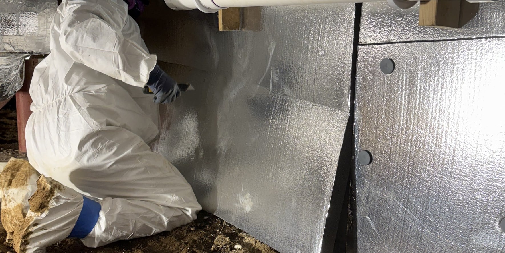 Indoor air quality in New Haven County CT — a PCRS technician in a protective suit fitting a foil vapour barrier against a crawlspace wall beneath the insulated trunk line